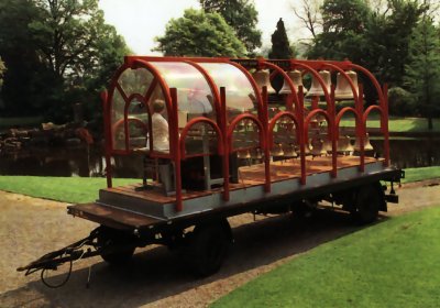 Dans un parc, une remorque de camion portant une structure où sont accrochées quelques dizaines de cloches de tailles différentes. Une personne est assise au clavier.